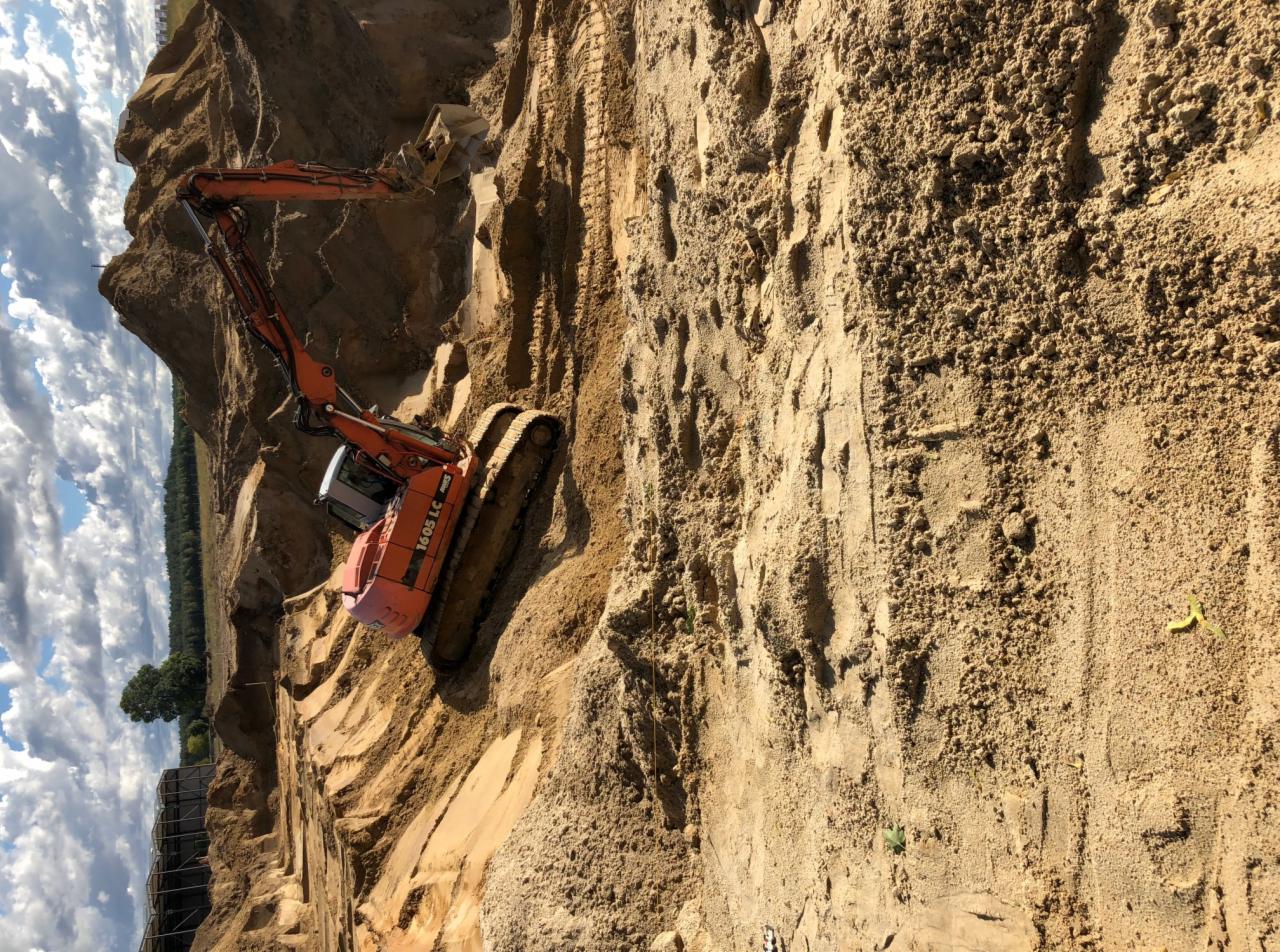 Pomarańczowa koparka gąsienicowa marki 1605 LC pracuje w wykopie z piasku, widoczne ślady gąsienic na ścianie wykopu, w tle drzewa i błękitne niebo z chmurami.