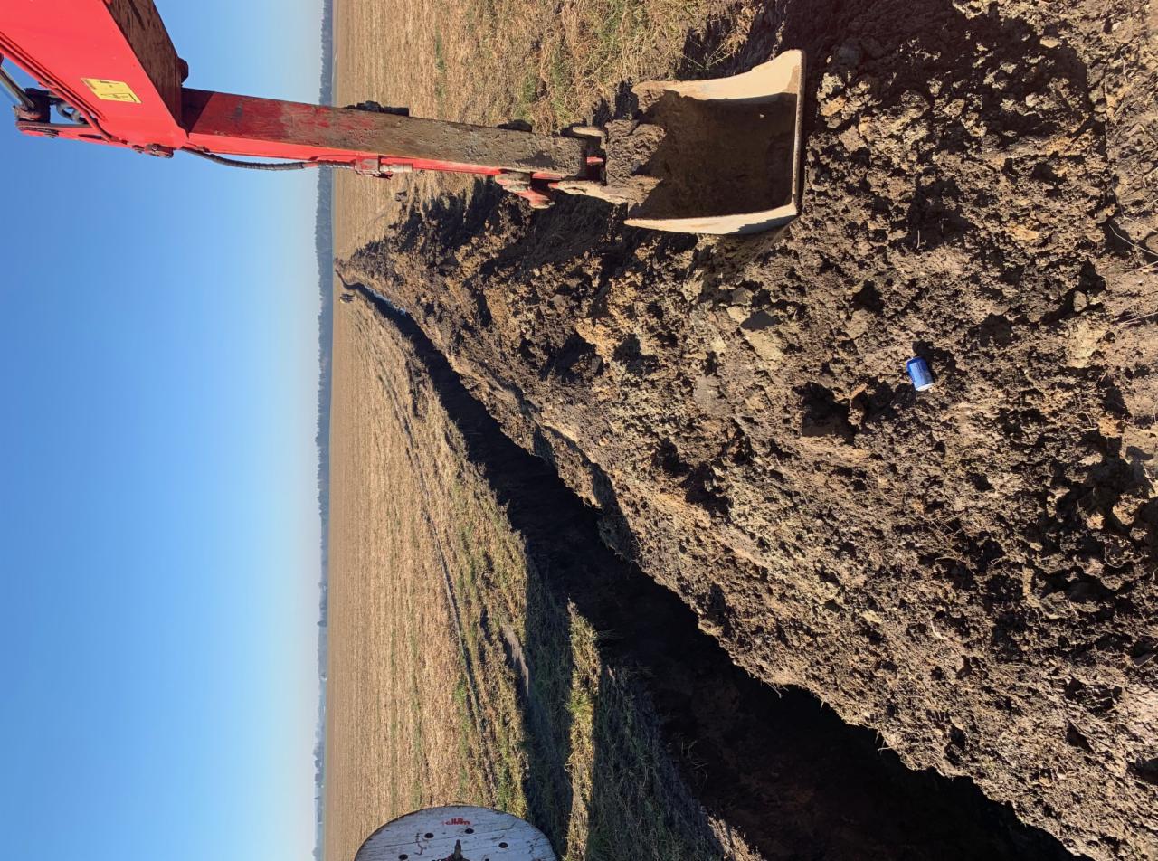 Czerwony koparka tworzy długi, wąski rów na polu, z widoczną szpulą kabla w rogu i puszką po napoju leżącą na ziemi.