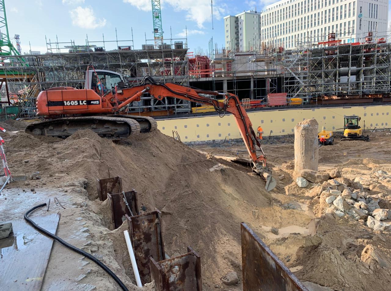 Pomarańczowa koparka gąsienicowa 1605 LC na placu budowy, w tle widoczne rusztowania, budynek w budowie oraz dwie mniejsze żółte koparki. W pierwszym planie stalowe ścianki szczelne.