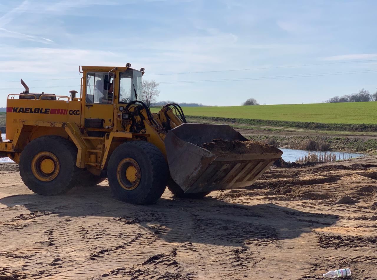 Żółta ładowarka Kaellble CKG z łyżką pełną ziemi na terenie budowy, w tle zielone pole i małe oczko wodne.