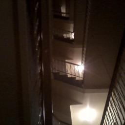 Jacoba - Wysokie schody w klatce schodowej budynku wielorodzinnego, widok z dołu, oświetlone punktowo, z metalową balustradą.