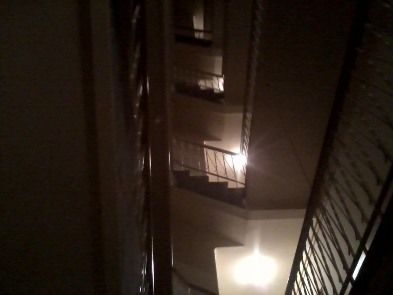 Wysokie schody w klatce schodowej budynku wielorodzinnego, widok z dołu, oświetlone punktowo, z metalową balustradą.