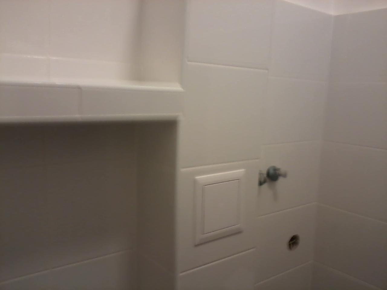 Białe, kwadratowe płytki ceramiczne na ścianie łazienki, z wnęką wyłożoną płytkami, mała klapka rewizyjna i wystający kran.