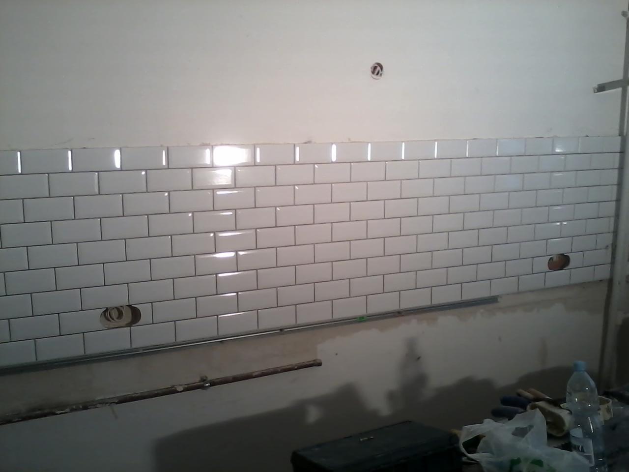 Ściana wyłożona białymi, błyszczącymi płytkami typu 'metro' z ciemną fugą, widoczne otwory na gniazdka elektryczne i narzędzia glazurnicze w dolnej części kadru.