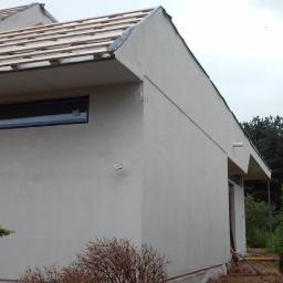 P.H.U. MAGIC ŁUKASZ GÓRKA - Boczny widok budynku po nałożeniu jasnego tynku elewacyjnego, z widocznymi elementami konstrukcji dachu i przygotowaniem pod montaż okien, otoczony zielenią.