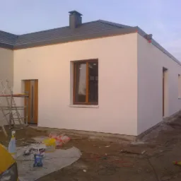 Świeżo otynkowany, biały budynek jednorodzinny z brązowymi ramami okiennymi i drzwiami, widoczne rusztowanie i akcesoria malarskie, teren wokół budynku jeszcze niezagospodarowany.