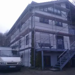 Trzypiętrowy dom w trakcie prac elewacyjnych, otoczony rusztowaniem, z zaparkowanym vanem dostawczym z przodu. Balustrada balkonu ze stali nierdzewnej. Widoczne drzewa bez liści w tle i pochmurne...