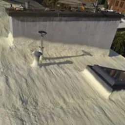 Poddasze po aplikacji izolacji natryskowej pianką, widoczna struktura piany na powierzchni, komin obłożony pianą, okno dachowe, ciepłe światło słoneczne.