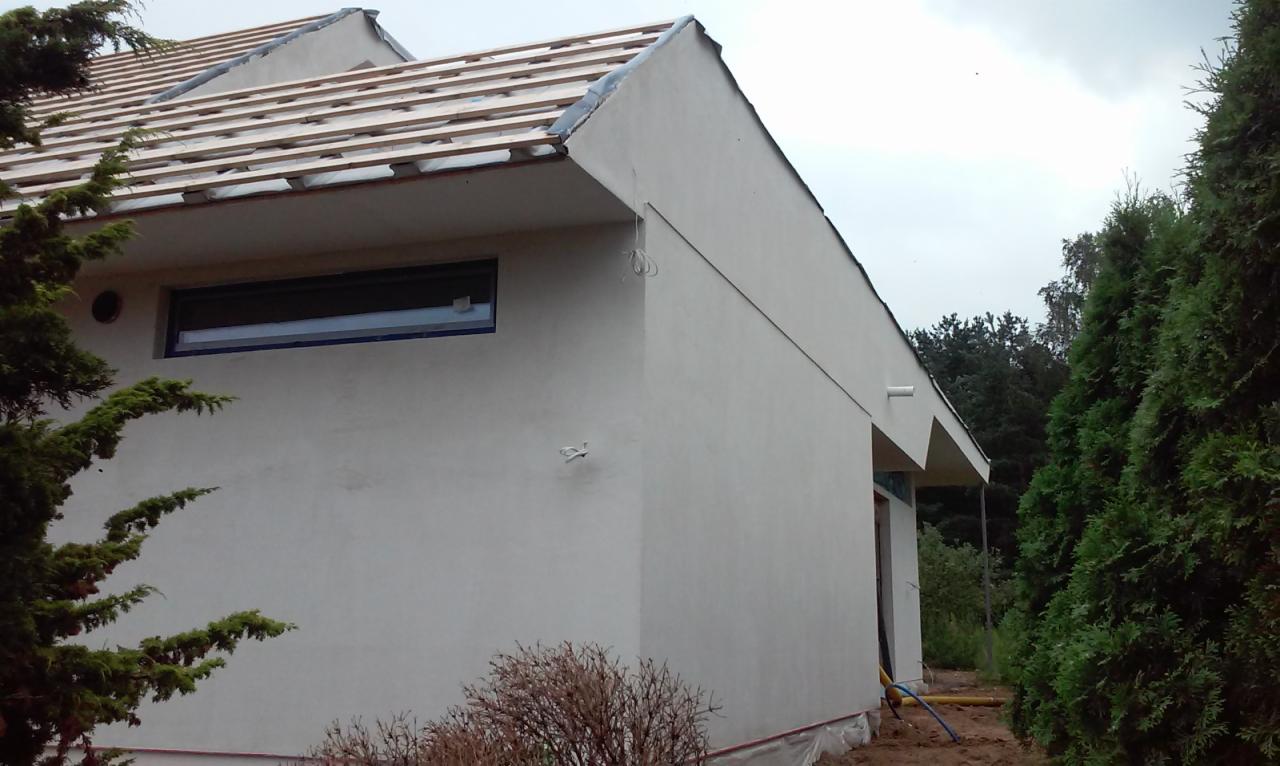 Boczny widok budynku po nałożeniu jasnego tynku elewacyjnego, z widocznymi elementami konstrukcji dachu i przygotowaniem pod montaż okien, otoczony zielenią.