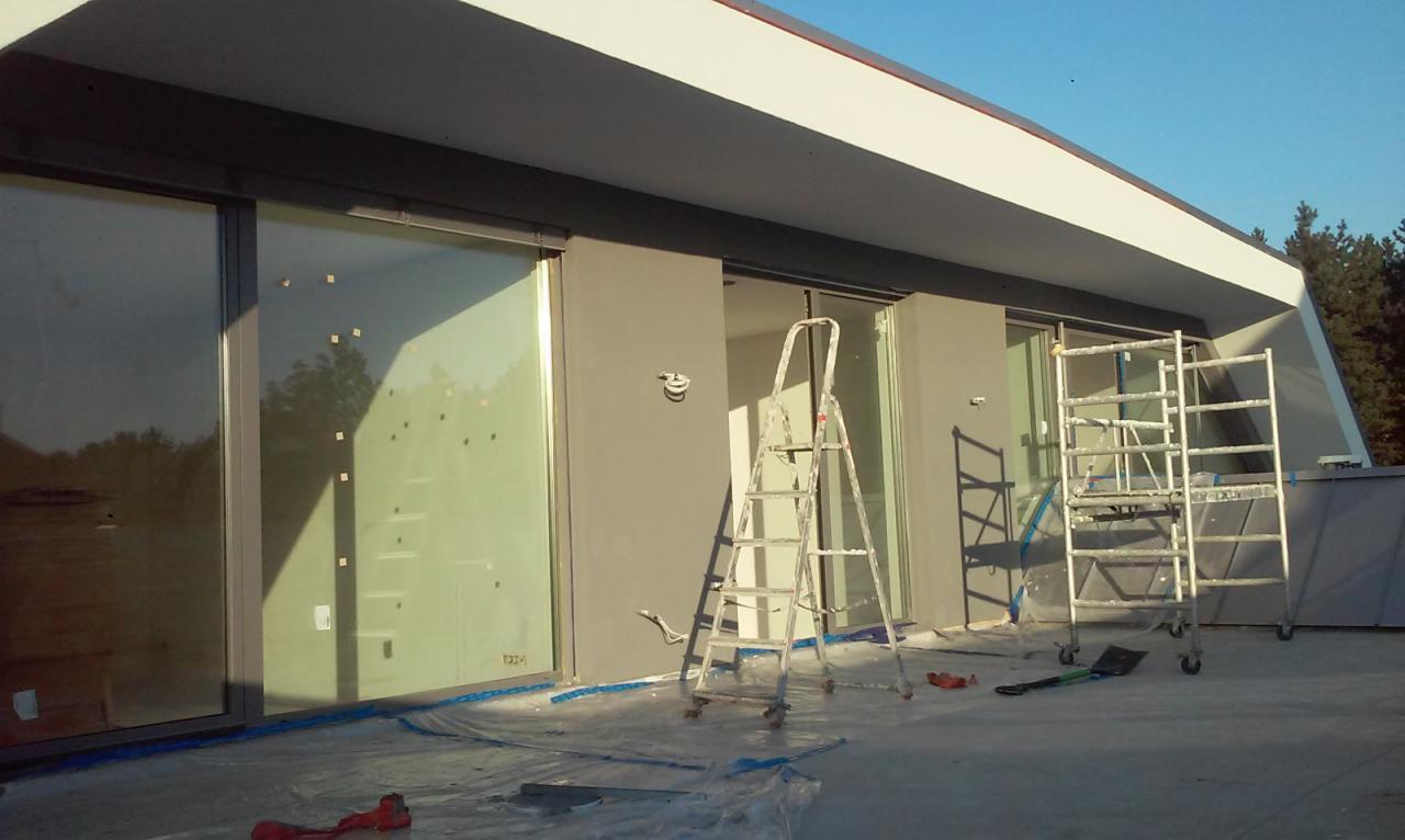 Świeżo pomalowana szara elewacja nowoczesnego domu z dużymi oknami, zabezpieczona folią malarską. Widoczne dwie drabiny, jedna przystawna i jedna rusztowanie, oraz narzędzia malarskie.