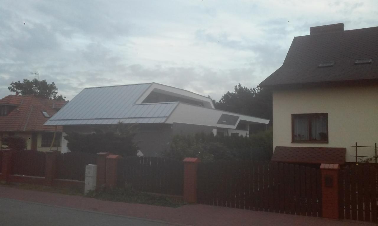 Nowoczesny dom z unikalnym dachem z blachy na tle pochmurnego nieba, widoczny zza drewnianego ogrodzenia.