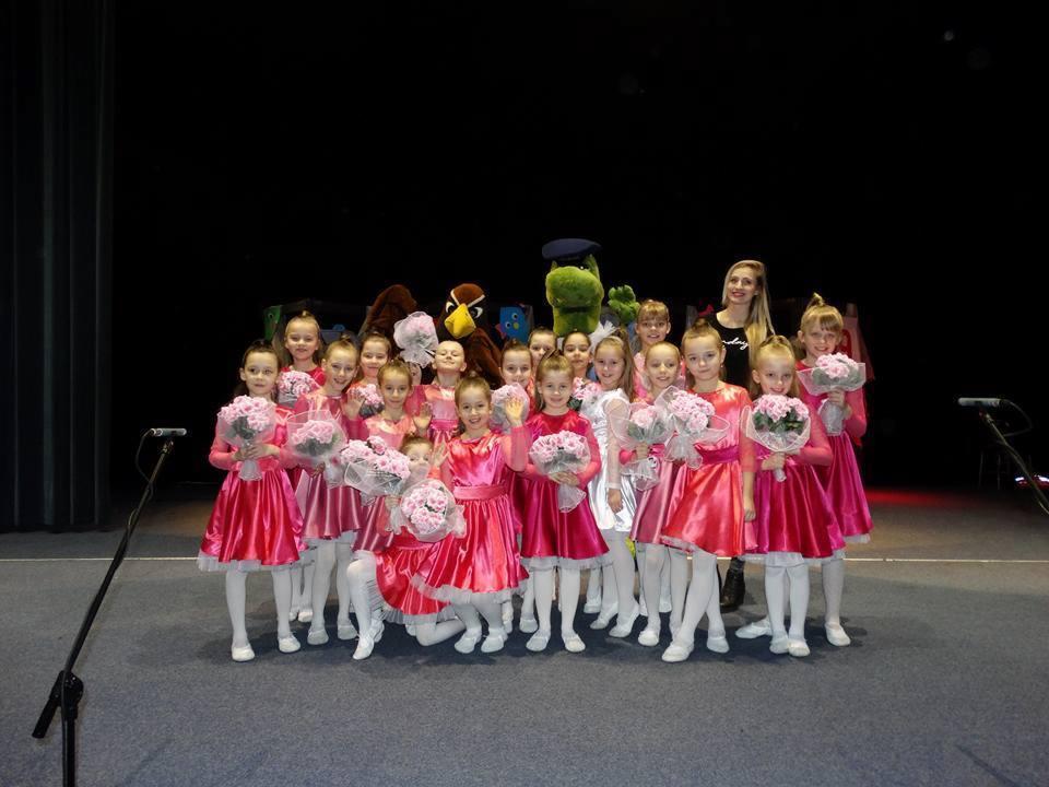 Grupa dziewczynek w błyszczących, różowych strojach tanecznych, z bukietami różowych kwiatów, pozuje na scenie z maskotkami