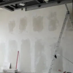 Wnętrze budynku w trakcie prac wykończeniowych, ściana z widocznymi łatami gipsowymi przed malowaniem, drabina oparta o ścianę, widoczne okno i elementy konstrukcyjne sufitu.