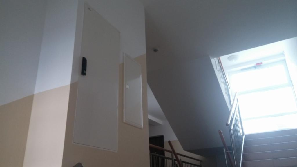 Białe szafki z zamkami na korytarzu, prawdopodobnie kryjące instalacje sieciowe. Widok na schody i okno z jasnym światłem dziennym.