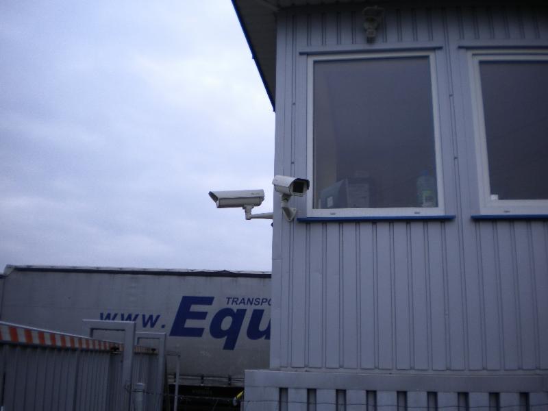 Dwie kamery CCTV zamontowane na szarym budynku z oknami, w tle widoczny fragment naczepy ciężarówki z napisem 'Transport'.
