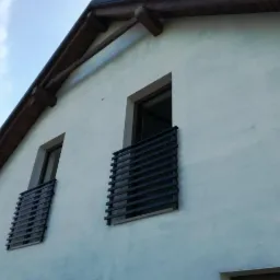 Dwa balkony z nowoczesnymi, poziomymi barierkami z ciemnego metalu na tle jasnej ściany domu z drewnianym okapem.