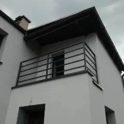 Szara, metalowa balustrada balkonowa z poziomymi prętami, zamontowana na białym budynku z widocznym oknem i ciemnym dachem w pochmurny dzień.