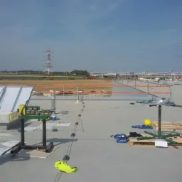 Montaż instalacji odgromowej na dachu hali przemysłowej, widoczne elementy systemu, przewody, uchwyty i narzędzia, w tle słupy wysokiego napięcia.