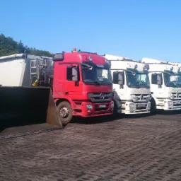 Żółta ładowarka Liebherr i rząd ciężarówek Mercedes na placu. W tle widać pryzmę materiału i drzewa. Słoneczny dzień.