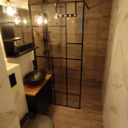 Nowoczesna łazienka z kabiną prysznicową w stylu loft, czarną armaturą i umywalką nablatową, oświetlona wiszącymi lampami z metalowymi kloszami.