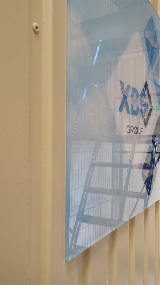 Logo 'XBS Group' na przezroczystej płycie, zamontowane na ścianie z widocznym odbiciem metalowej konstrukcji w tle.