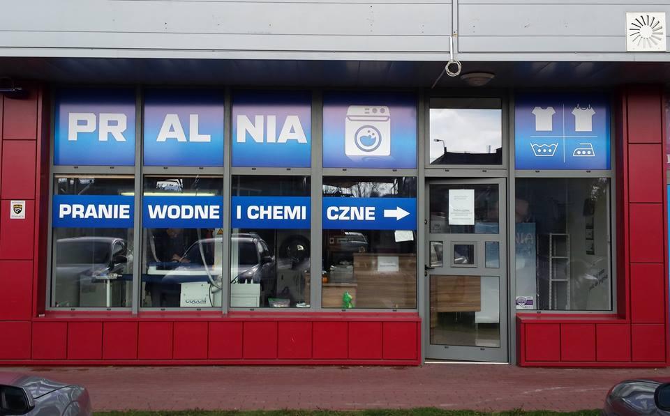Fasada pralni chemicznej z niebieskimi grafikami na oknach, w tym logo pralki i napisy 'PRALNIA' oraz 'PRANIE WODNE I CHEMICZNE'. Widoczne wnętrze z urządzeniami.