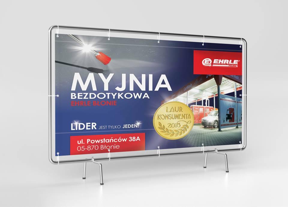 Stojak reklamowy myjni bezdotykowej Ehrle Błonie z nagrodą Laur Konsumenta 2015, prezentujący adres ul. Powstańców 38A. Grafika w niebiesko-szaro-czerwonej kolorystyce.