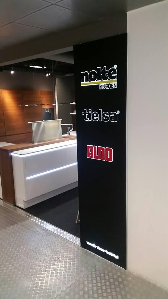 Wnętrze salonu meblowego z ekspozycją kuchni. Czarny panel z logo marek: Nokte Kuchen, Tielsa, Alno. Nowoczesna zabudowa kuchenna w tle.