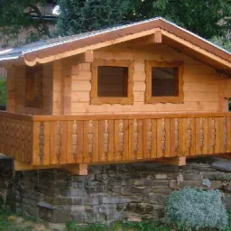 Mały, parterowy domek z bali z balkonem, osadzony na kamiennej konstrukcji. Drewniane wykończenia okien i balustrady. W tle zieleń drzew.