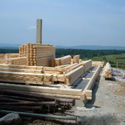 Budowa domu z bali: ułożone drewniane elementy konstrukcyjne na fundamencie, w tle zielone wzgórza i błękitne niebo. Widoczne podpory i narzędzia.