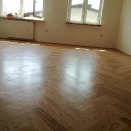 Parkiet ułożony w jodełkę w jasnym, pustym pokoju z dwoma oknami, grzejnikiem i widocznymi śladami po gniazdkach elektrycznych na ścianach.