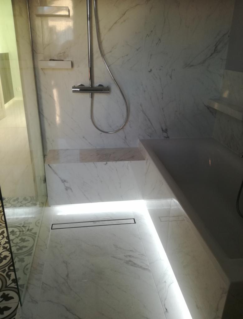 Nowoczesna kabina prysznicowa wykończona białym marmurem z subtelnym podświetleniem LED pod półką i wzdłuż ściany, z odpływem liniowym w podłodze i minimalistyczną armaturą.