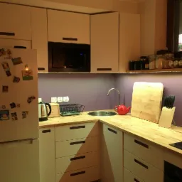 Jasna kuchnia z drewnianym blatem, fioletową ścianą i białymi szafkami. Na blacie czajnik, deska do krojenia i pojemnik z nożami. Na lodówce magnesy.