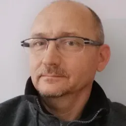 Portret mężczyzny w okularach i szarej bluzie na tle jasnej ściany.