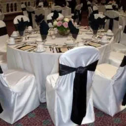 Uroczysta sala bankietowa z okrągłymi stołami nakrytymi białymi obrusami, krzesła w białych pokrowcach z czarnymi szarfami, centralnie umieszczone kompozycje kwiatowe w szklanych wazonach.