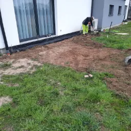 Prace ziemne przy nowym domu: robotnik w jaskrawożółtych spodniach kopie łopatą obok wykopu zabezpieczonego czarną folią drenażową, w tle minikoparka i sterta cegieł.