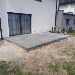 Ułożony taras z szarych płyt betonowych przylegający do nowoczesnego, białego domu z dużym oknem przesuwnym, teren wokół jeszcze niezagospodarowany.