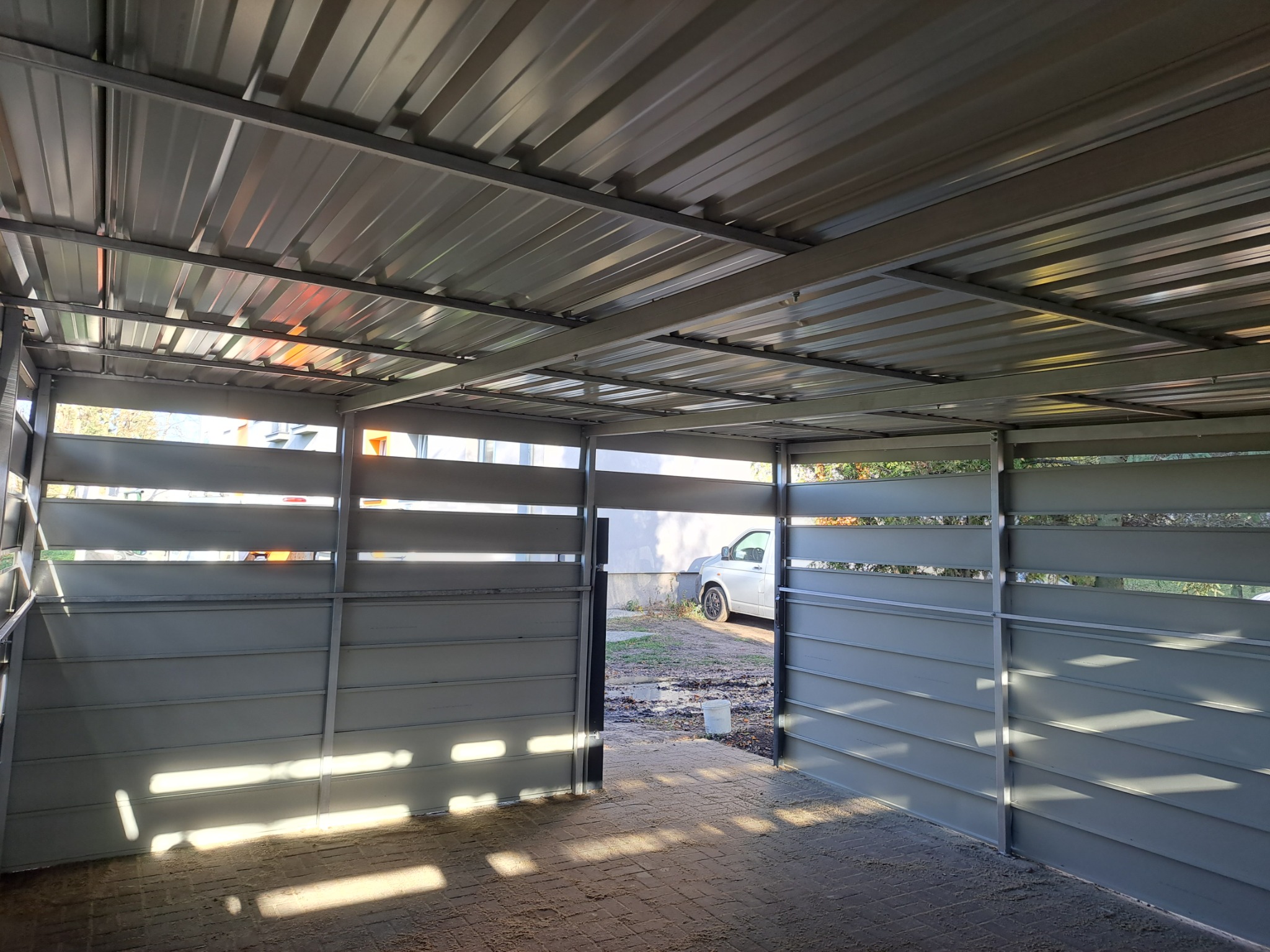 Zadaszona wiata garażowa z szarej blachy trapezowej, widok od wewnątrz na kostkę brukową i zaparkowany samochód. Konstrukcja stalowa z poziomymi prześwitami.