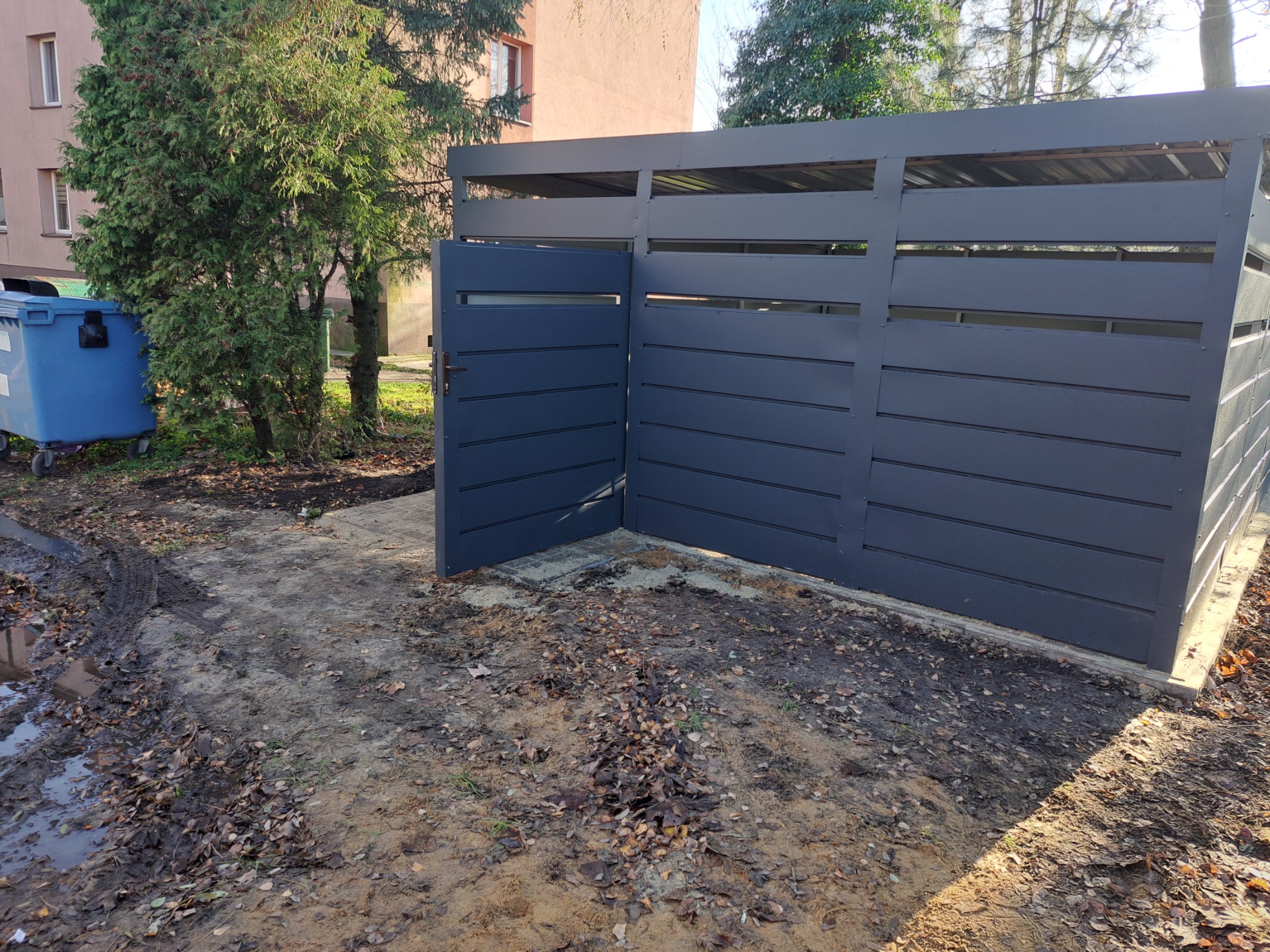 Nowoczesna, szara wiata śmietnikowa z otwartymi drzwiami, otoczona ziemią i liśćmi. W tle budynek mieszkalny i drzewa.