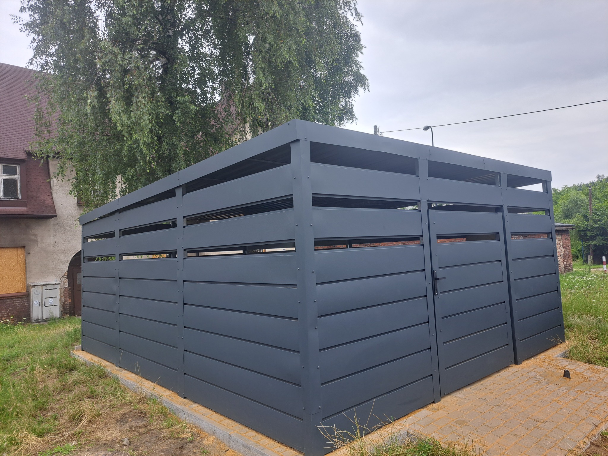 Nowoczesna, grafitowa obudowa śmietnikowa z poziomych paneli, otoczona kostką brukową, na tle budynku z cegły i zieleni. Widok z perspektywy.