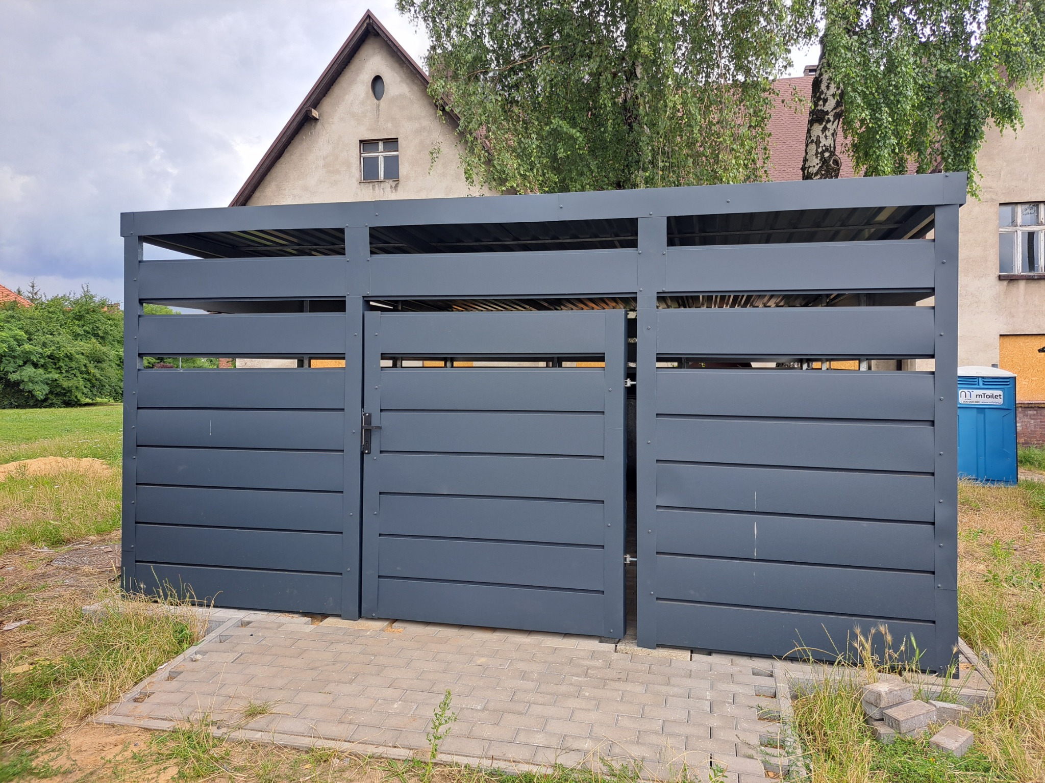 Nowoczesna, szara wiata śmietnikowa z poziomymi panelami, częściowo otwarta, na kostce brukowej. W tle budynek mieszkalny i przenośna toaleta.