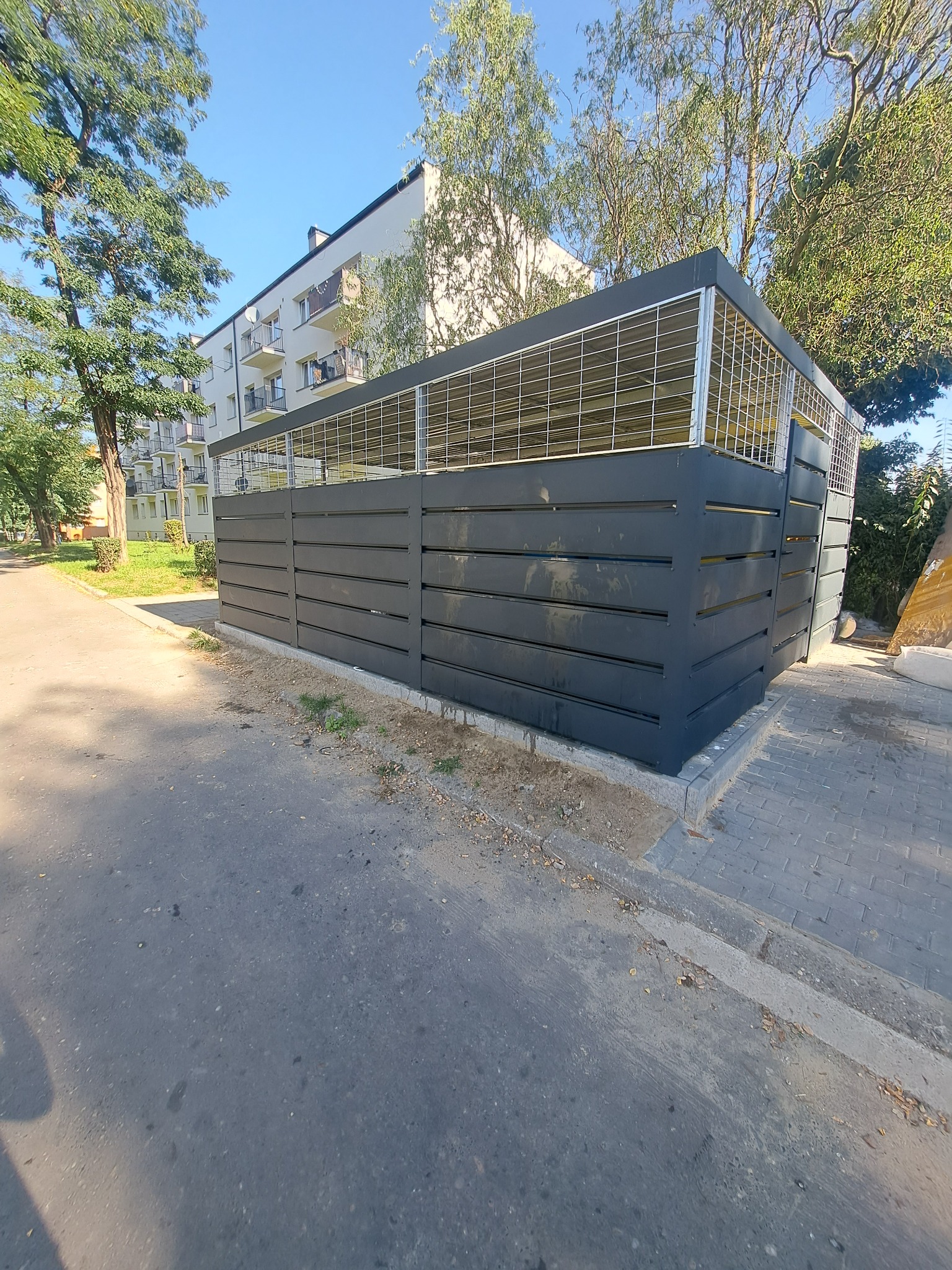 Nowoczesna zabudowa śmietnikowa z szarego drewna i metalowej siatki, na tle bloku mieszkalnego i zieleni. Widok z poziomu chodnika, w słoneczny dzień.