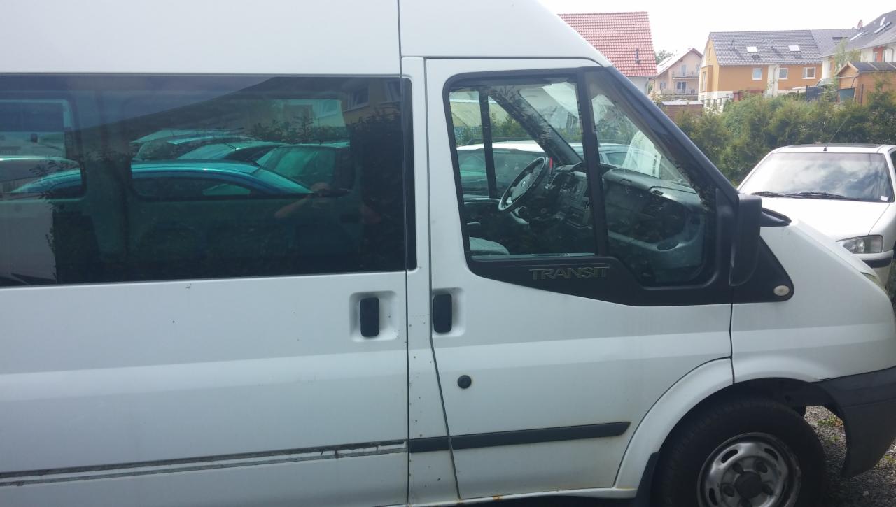 Biały bus marki Transit z widocznymi szybami, zarysami wnętrza i innymi zaparkowanymi pojazdami w tle.