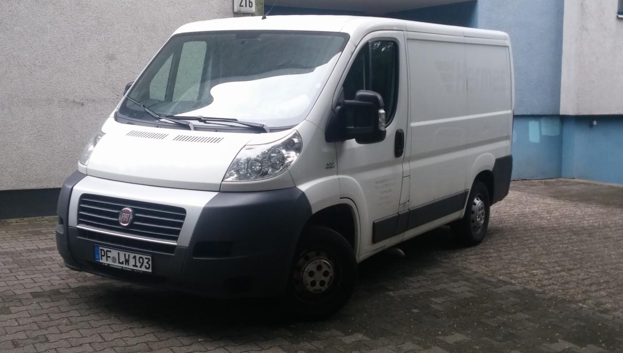 Biały furgon Fiat Ducato z niemiecką tablicą rejestracyjną PF LW 193, zaparkowany na brukowanym podwórzu przed budynkiem.