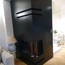 Nowoczesny, czarny kominek narożny z geometrycznymi zdobieniami, widoczny w salonie z parkietem i elementami wystroju w stonowanych kolorach.