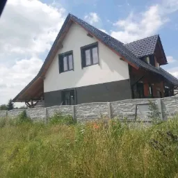 Nowoczesny dom jednorodzinny z szarą elewacją w dolnej części i białą w górnej, grafitowa dachówka, ogrodzony betonowym płotem, widok zza wysokiej trawy.