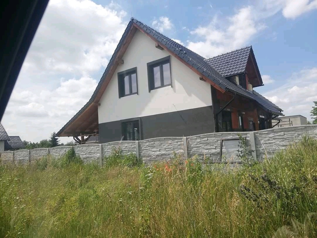 Nowoczesny dom jednorodzinny z szarą elewacją w dolnej części i białą w górnej, grafitowa dachówka, ogrodzony betonowym płotem, widok zza wysokiej trawy.