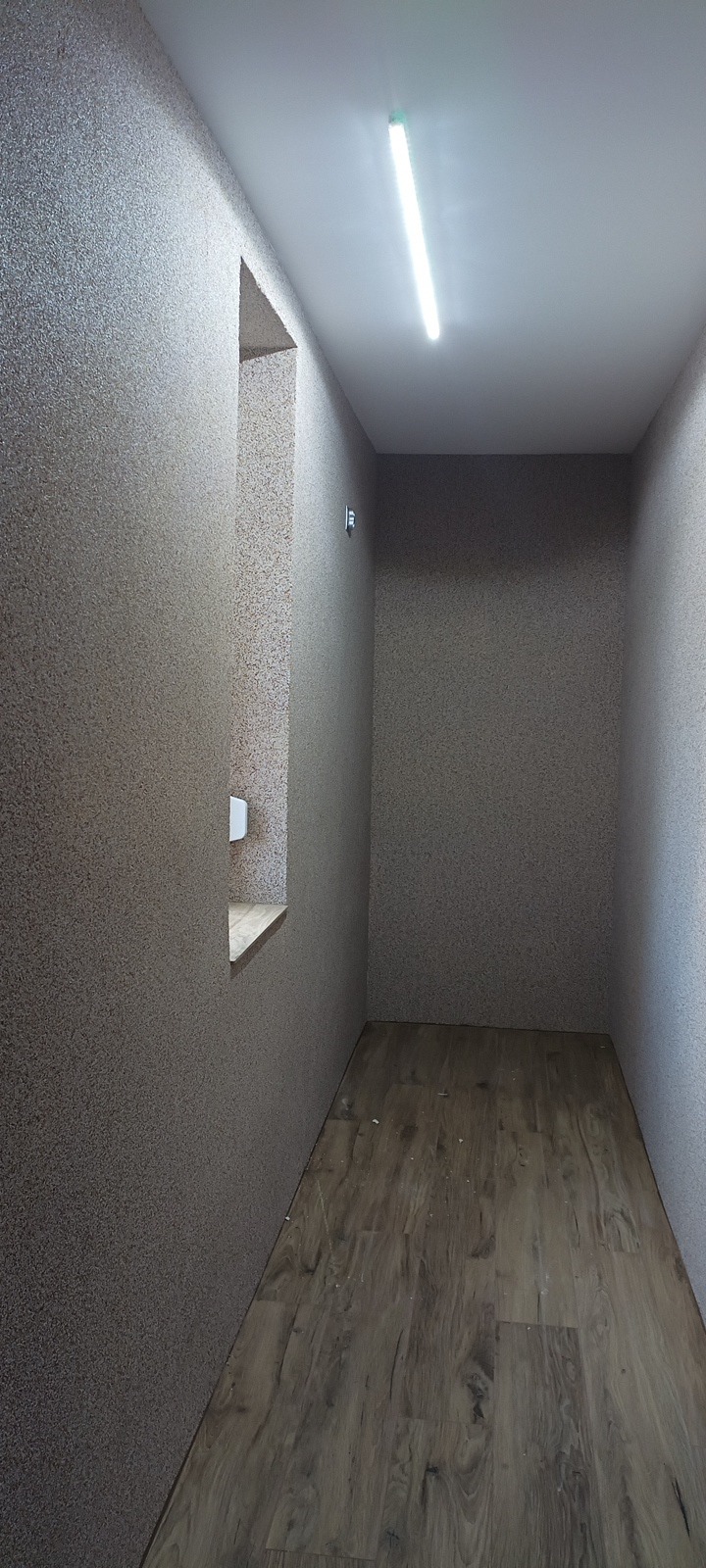 Wnętrze małego pomieszczenia z teksturowanymi ścianami w odcieniach beżu i brązu, podłoga wyłożona panelami imitującymi drewno, oświetlone pojedynczą lampą jarzeniową na suficie i wąskim oknem...