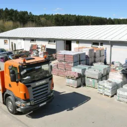 Pomarańczowa ciężarówka Scania z dźwigiem na tle magazynu ze stertami materiałów budowlanych, w tym kostki i cegieł, w słoneczny dzień.