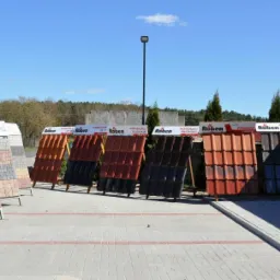 Ekspozycja różnorodnych materiałów budowlanych: kostki betonowe i dachówki RÖBEN, KORAMIC, prezentowanych na stojakach przed budynkiem w słoneczny dzień.